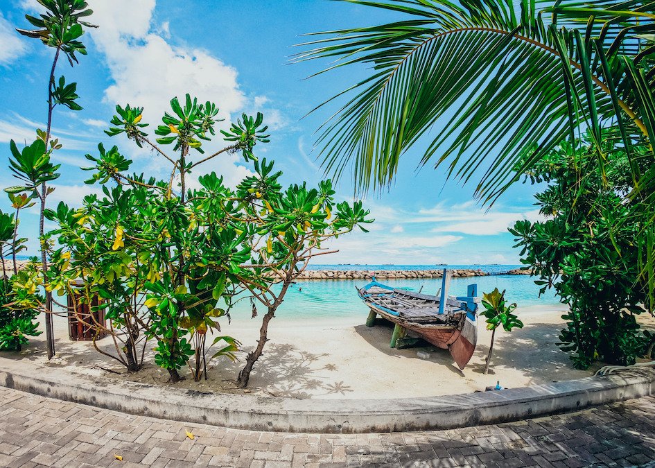 A scenic shot of a beach in the Maldives, showing lush greenery, white sand, the bright blue sea and a boat on the sand.